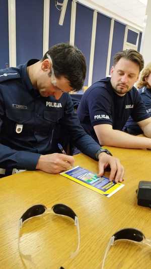 Funkcjonariusz polskiej Policji siedząc za stołem próbuje złożyć podpis na jego oczach okulary symulujące chorobę wzroku