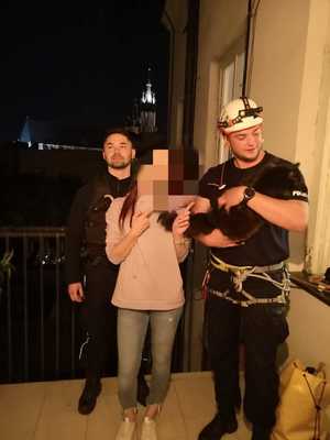 policjant trzyma na ręku uratowanego kota, obok stoi kobieta, która ma zamazana twarz, a z tyłu drugi funkcjonariusz