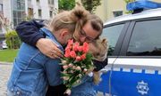 Mama - policjantka w mundurze trzyma w objęciach dwie dziewczynki, a w dłoni bukiet kwiatów. Obok zaparkowany jest policyjny radiowóz