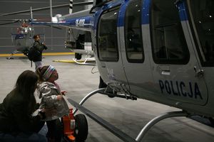 Przed policyjnym śmigłowcem Bell w hangarze stoi kilkuletnia dziewczynka, patrząca na maszynę. Stojące dziecko stabilizuje opiekunka, podtrzymując dziecko za biodra.