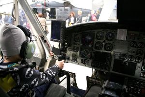Zdjęcie wykonane z przedziału pasażersko-transportowego śmigłowca. Za wolantem na miejscu pilota siedzi kilkuletni chłopiec ubrany w czarną kurtkę z białymi grafikami Batmana.