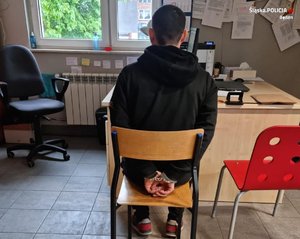 Zatrzymany mężczyzna, który ma założone kajdanki na ręce trzymane z tyłu, siedzący w jednym z pokoi na komisariacie