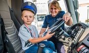 Policjantka  stoi przy otwartym radiowozie, a dziecko w policyjnej czapce siedzi za kierownicą auta