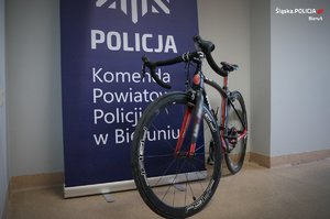 Przy granatowym banerze, na którym widać białe napisy: POLICJA i Komenda Powiatowa Policji w Bieruniu, stoi zabezpieczony przez policjantów rower