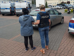 Policjantka prowadzi zatrzymaną kobietę. Policjantka ma na sobie czarną kamizelkę z białym napisem Policja na plecach