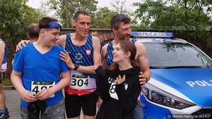 Dwójka uśmiechniętych niepełnosprawnych młodych biegaczy w asyście dwóch policyjnych lekkoatletów stojących przy policyjnym radiowozie