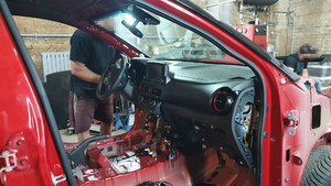 Zdjęcie przedstawia wnętrze garażu, a w nim częściowo zdemontowany czerwony pojazd. W tle widać dwie osoby, ich twarze są zasłonięte
