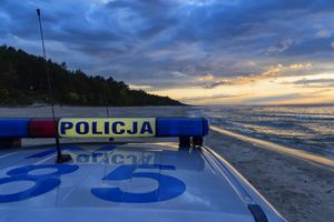 Dach policyjnego radiowozu z sygnałami wizualnymi i napisem Policja, w tle plaża i morze.
