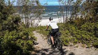 Mężczyzna jedzie rowerem przez wydmę w stronę nadmorskiej plaży. Ubrany jest w białą koszulkę z napisem Policja, krótkie spodenki i kask rowerowy. W tle widać morze.