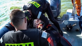 Dwóch mężczyzn ubranych w ubrania do pływania z pianki znajduje się w pontonie z silnikiem motorowym. Maja na sobie kamizelki z napisem Policja.