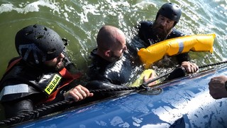 Trzech mężczyzn ubranych w czarne kombinezony pływackie z pianki zanurzonych jest w wodzie. Na ubraniu jednego z nich widoczny napis: Policja. Mężczyźni trzymają się liny przymocowanej do pontonu.