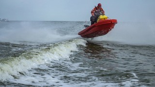 Mężczyzna na skuterze wodnym płynie z dużą prędkością po wodzie. Ubrany jest w specjalistyczny strój ratowniczy oraz kask. Skuter unosi się wysoko na falach.