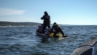Mężczyzna w stroju pływackim z pianki z napisem na plecach Policja stoi na skuterze na środku morza. Drugi, ubrany tak jak on, klęczy na pontonie przymocowanym do skutera. Patrzą w stronę plaży.