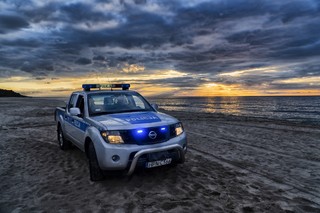 Oznakowany samochód policyjny zaparkowany na nadmorskiej plaży, sygnalizacja uprzywilejowania pojazdu jest włączona, w tle fragment lasu i zachodzącego słońca.