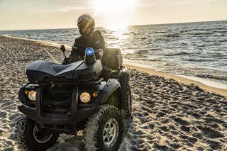 Mężczyzna w kasku jedzie czterokołowym pojazdem terenowym po nadmorskiej plaży. Z boku pojazdu znajduje włączony uprzywilejowany znak sygnalizacyjny. Promienie słoneczne odbijają się w wodzie.