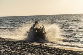 Mężczyzna jedzie wzdłuż brzegu morza czterokołowym pojazdem terenowym. Woda rozpryskuje się na boki.