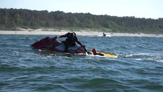Mężczyzna w stroju pływackim z pianki siedzi na skuterze wodnym na środku morza. Podaje rękę osobie znajdującej się w wodzie i trzymającej się doczepionego do skutera materaca ratowniczego z licznymi uchwytami.