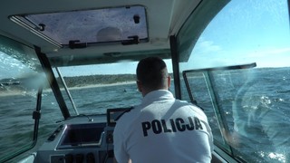 Wnętrze łodzi patrolującej, za sterami której siedzi policjant. Łódź płynie w kierunku plaży.