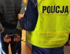 Mężczyzna w kamizelce odblaskowej z napisem policja trzyma innego mężczyznę pod ramię, który ma kajdanki na rękach