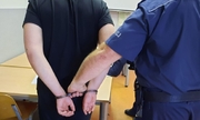 Policjant w mundurze trzyma za kajdanki, które są założone innemu mężczyźnie na ręce trzymane z tyłu