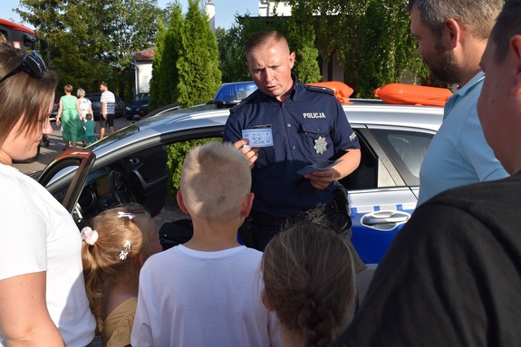 Policjant przy radiowozie rozdaje ulotki grupie dzieci.