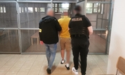 Dwóch policjantów w ubraniach cywilnych, jeden w kamizelce, a drugi z opaską na ręku z napisem policja prowadzą podejrzanego, który ma założone kajdanki zespolone - na ręce i nogi