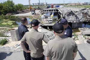 dwaj umundurowani funkcjonariusze i dwaj mężczyźni w zielonych koszulkach polo oglądają skalę zniszczeń wywołanych atakiem Rosji na Ukrainę