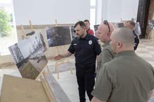 dwaj umundurowani funkcjonariusze i dwaj mężczyźni w zielonych koszulkach polo oglądają skalę zniszczeń i zbrodni wywołanych atakiem Rosji na Ukrainę, które udokumentowane są na zdjęciach ustawionych na sztalugach