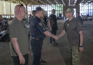 umundurowani funkcjonariusze i mężczyźni w zielonych koszulkach polo w hali w której znajduje się zniszczony sprzęt w wyniku ataku Rosji na Ukrainę
