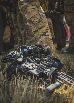 Na pierwszym planie w trawie pistolet maszynowy z celownikiem optycznym w tle nogi kontrterorystów w mundurach ćwiczebnych.