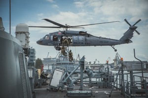 Fotografia wykonana przez instalacje pokładowe okrętu, na który desantują się policyjni kontrterroryści. Policyjny Black Hawk w zawisie nad lądowiskiem śmigłowcowym usytuowanym na rufie okrętu Marynarki Wojennej RP.