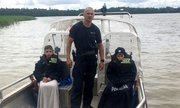 Policjant stoi na pokładzie łodzi, a po jego obu stronach siedzą młodzi chłopcy przykryci policyjnymi mundurami