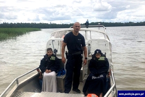 Policjant stoi na pokładzie łodzi, a po jego obu stronach siedzą młodzi chłopcy przykryci policyjnymi mundurami