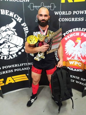 Starszy Posterunkowy Grzegorz Rólka w stroju spotowym podczas mistrzostw, trzyma 2 puchary, dyplomy, w lewej ręce plecak