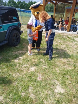 Policjantka daje dziecku worek odblaskowy promujący akcję &quot;Kręci mnie bezpieczeństwo nad wodą&quot;, obok stoi maskotka policyjna i samochód służbowy straży granicznej.