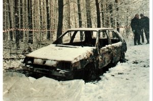 stojący w lesie wrak spalonego samochodu. za nim widać ludzi