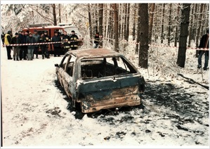 stojący w lesie wrak spalonego samochodu. w głębi widać grupę ludzi