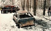 stojący w lesie wrak spalonego samochodu. w głębi widać grupę ludzi
