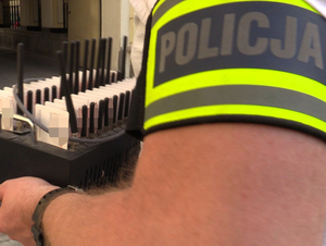 na pierwszym planie widoczna opaska z napisem Policja na ramieniu funkcjonariusza, a w tle zabezpieczony sprzęt informatyczny