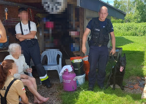 policyjny przewodnik z psem odnaleziona kobieta siedząca na krześle obok niej stojący strażacy na zewnątrz przed szopą
