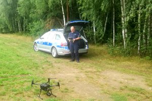policjant w mundurze przy radiowozie i sterującym dronem z ziemi, w oddali las