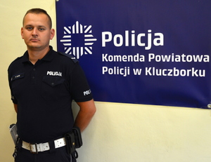 policjant stoi na tle ściany, na ścianie wisi banner z napisem Policja Komenda Powiatowa Policji w Kluczborku