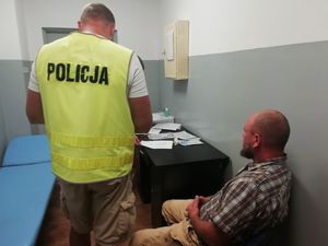 Mężczyzna siedzący przy biurku, obok stojący policjant w kamizelce z napisem Policja