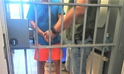 Zdjęcie przedstawia zatrzymanego mężczyznę stojącego za kratą pomieszczenia dla osób zatrzymanych. Obok niego znajduje się funkcjonariusz