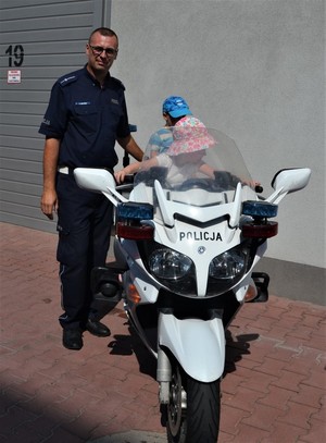 Dwoje dzieci siedzi na motocyklu policyjnym obok stoi policjant.