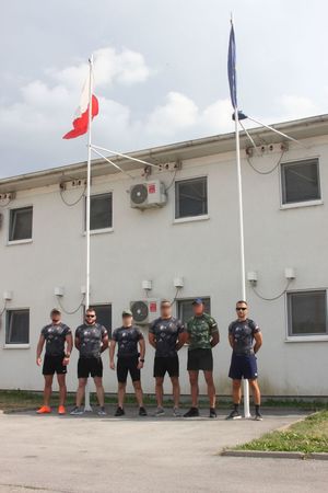 sześciu funkcjonariuszy XXXIII Rotacji Jednostki Specjalnej Polskiej Policji w Kosowie w strojach sportowych stoi pod masztem z flagami Polski i Unii Europejskiej. W tle widać budynek