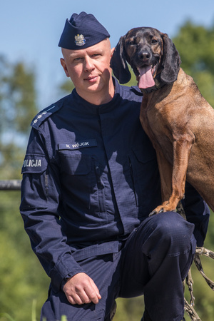 Policjant przyklęka na jedno kolano, obok niego stoi jego pies policyjny.