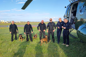 Czterej policjanci, trzy psy służbowe i dwaj policyjni lotnicy obok śmigłowca na trawiastym lądowisku.