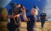 policjant trzyma na rękach psa policyjnego, policjantka stojąca obok głaszcze psa, w tle idący policjant i balon w trakcie napełniania powietrzem