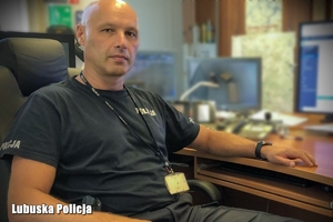 oficer dyżurny w koszulce  napisem Policja siedzi bokiem do biurka, przodem do obiektywu